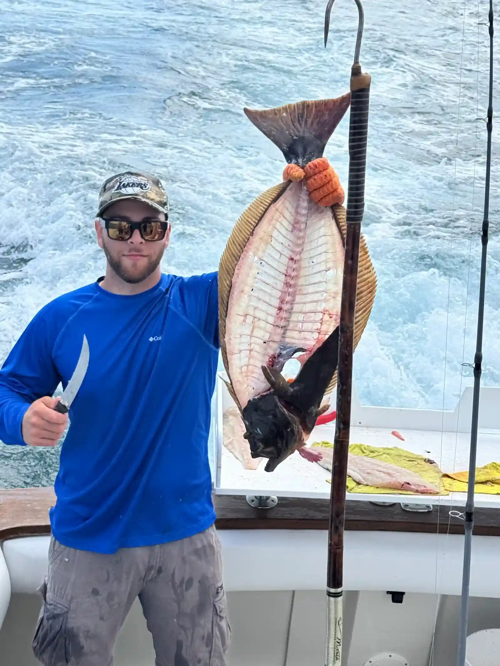 Angler holding a fillet knife beside a halibut hanging from a gaff on the deck of the Mystic Queen